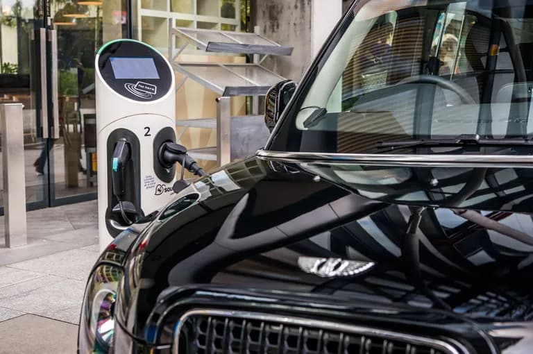 A black car plugged into a white electric vehicle charging station on a city sidewalk.